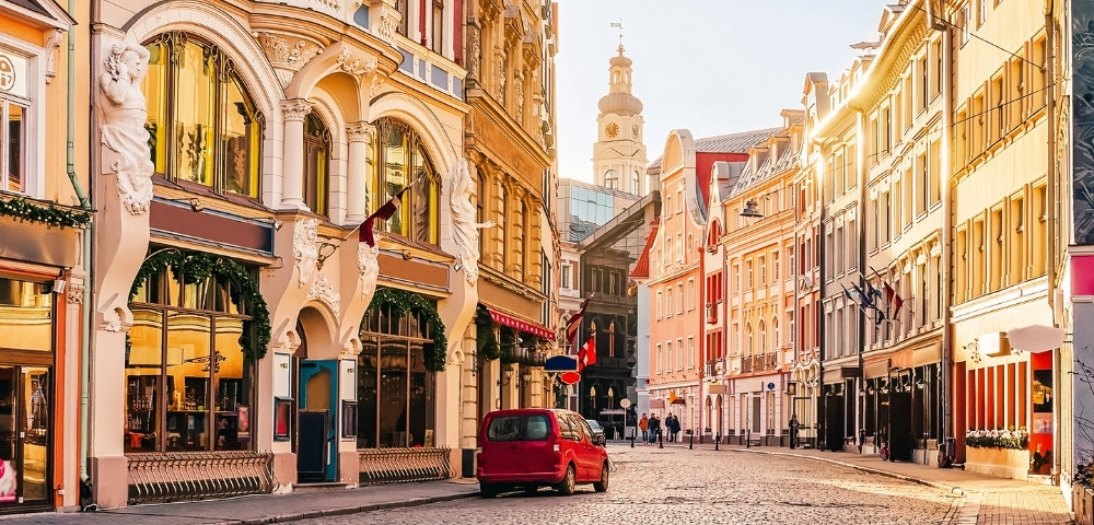 Charming European street with colorful historic buildings illuminated by warm sunlight. A red car and a few pedestrians on the cobblestone road convey a peaceful morning atmosphere.