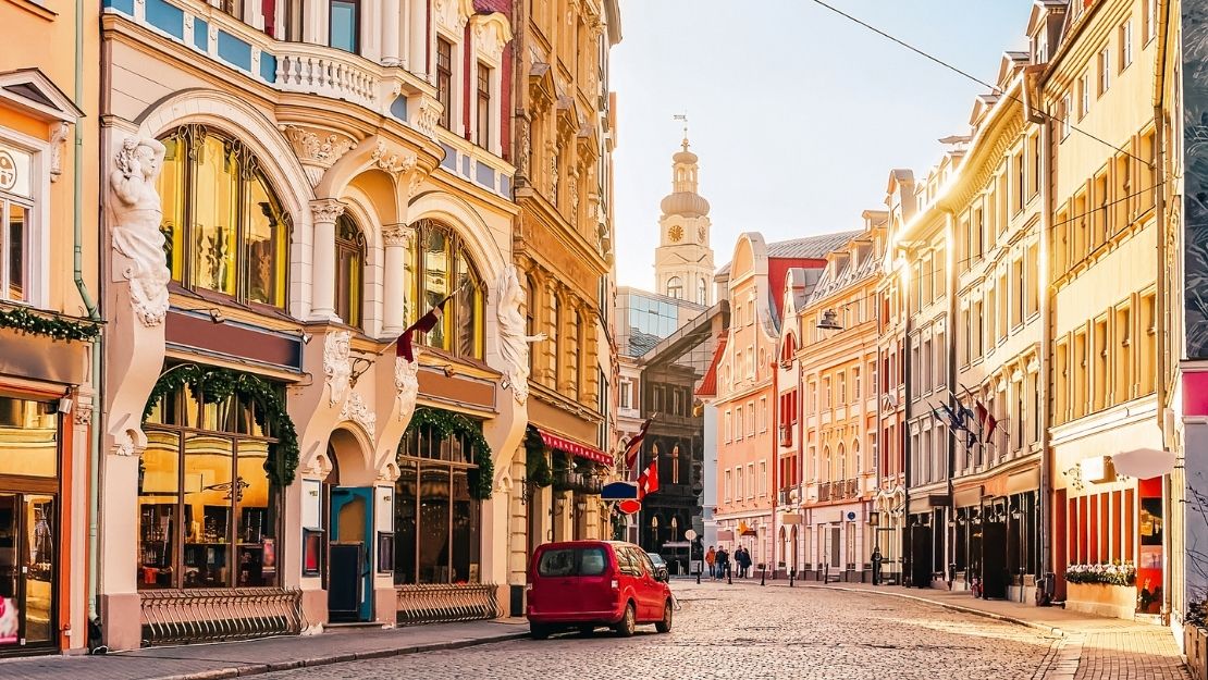 Charming European street with colorful historic buildings illuminated by warm sunlight. A red car and a few pedestrians on the cobblestone road convey a peaceful morning atmosphere.