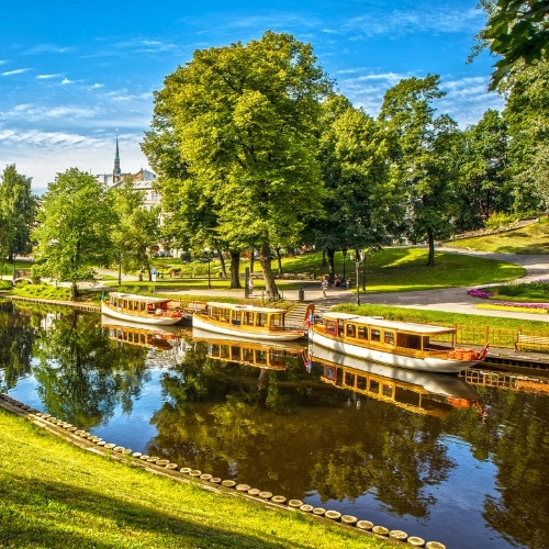 Sunlit park scene with a serene river reflecting lush green trees. Three small, docked boats sit along a grassy bank, under a bright, blue sky.