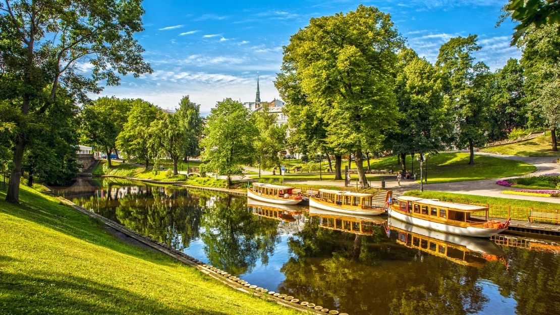 Sunlit park scene with a serene river reflecting lush green trees. Three small, docked boats sit along a grassy bank, under a bright, blue sky.