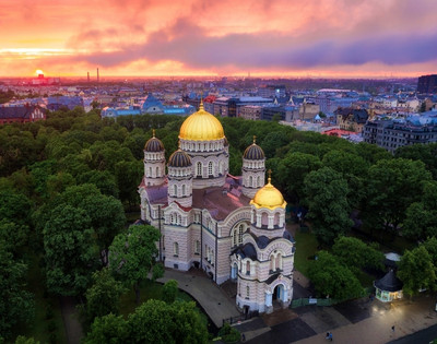 Aerial view of a grand cathedral with golden domes surrounded by lush green trees, set against a cityscape at sunset, casting a warm, serene glow.