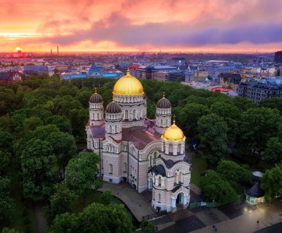 Aerial view of a grand cathedral with golden domes surrounded by lush green trees, set against a cityscape at sunset, casting a warm, serene glow.