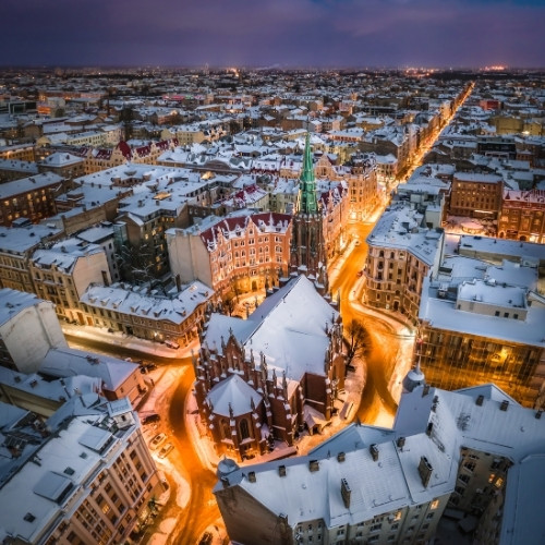 Aerial view of a snow-covered cityscape featuring historic architecture, glowing warmly in streetlights. Central church, pointed spire; cozy, serene ambiance.