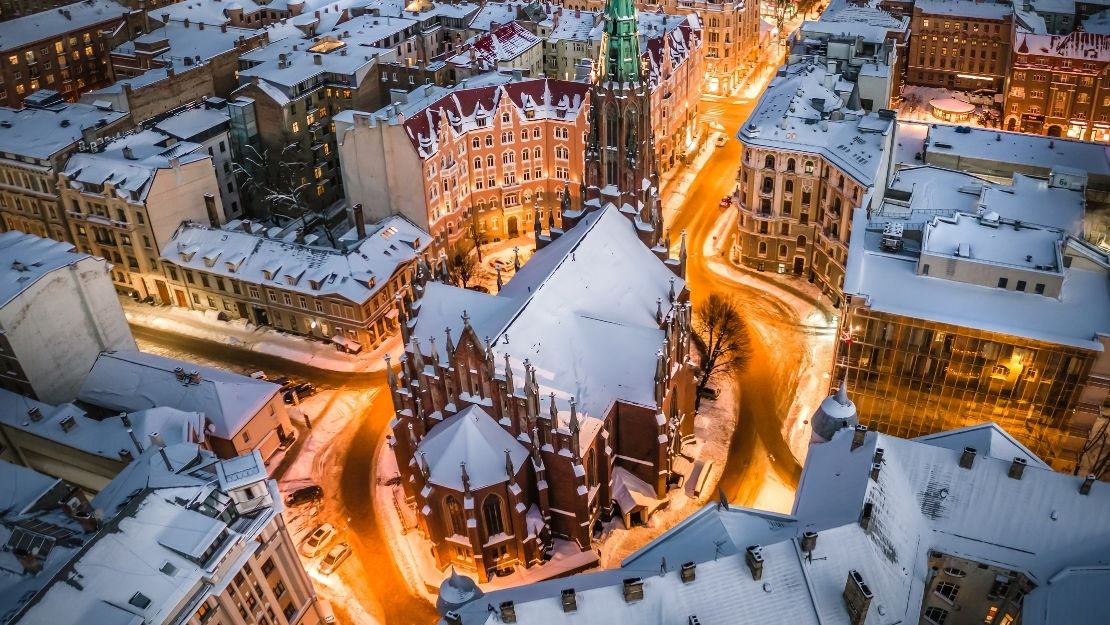 Aerial view of a snow-covered cityscape featuring historic architecture, glowing warmly in streetlights.
