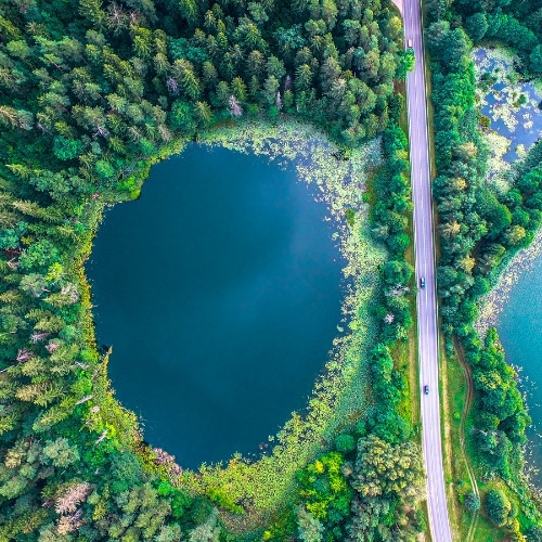 Aerial view of two adjacent lakes surrounded by dense green forest. A road runs vertically between them, with a few cars visible.