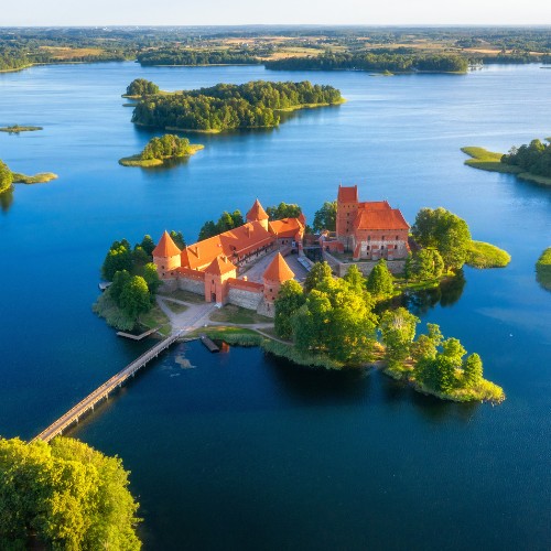 Aerial view of a red-roofed castle on an island, surrounded by blue lake waters and lush greenery, connected by a bridge, under a clear sky.