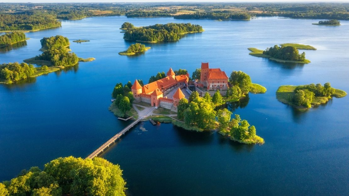 Aerial view of a red-roofed castle on an island, surrounded by blue lake waters and lush greenery, connected by a bridge, under a clear sky.