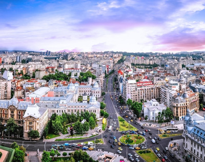 Aerial view of a bustling cityscape at dusk, featuring historic and modern buildings, busy streets, and green spaces, under a vibrant purple and pink sky.