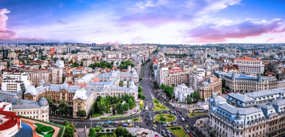 Aerial view of a bustling cityscape at dusk, featuring historic and modern buildings, busy streets, and green spaces, under a vibrant purple and pink sky.
