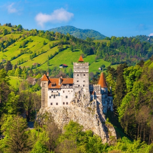 Fairytale-like castle with red roofs perched on a rocky hill, surrounded by lush green forests and rolling hills under a bright blue sky.
