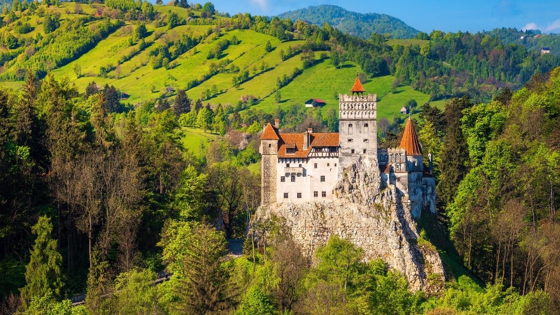 Fairytale-like castle with red roofs perched on a rocky hill, surrounded by lush green forests and rolling hills under a bright blue sky.