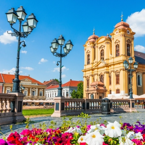 A vibrant European square with a baroque church adorned in warm yellow hues, surrounded by historic buildings. Foreground features colorful flowers and ornate lampposts under a blue sky.