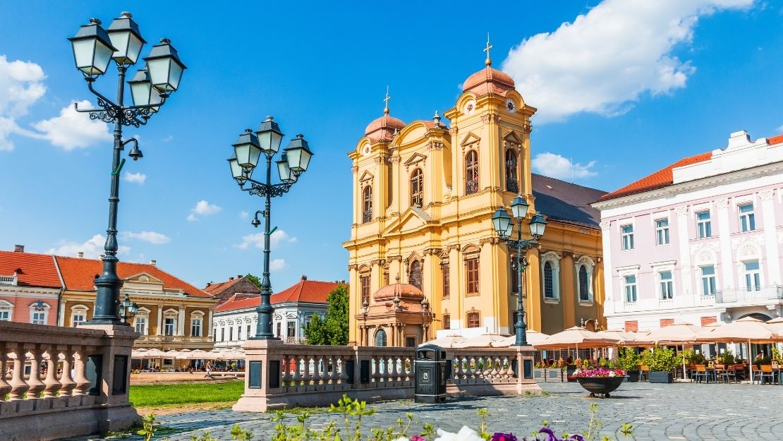 A vibrant European square with a baroque church adorned in warm yellow hues, surrounded by historic buildings. Foreground features colorful flowers and ornate lampposts under a blue sky.