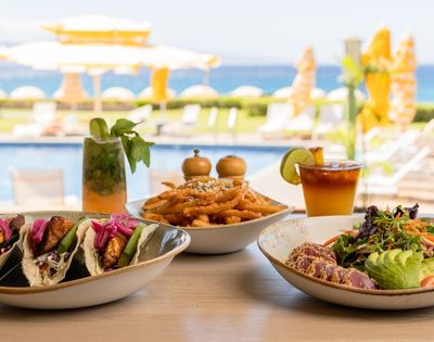 A table with fish tacos, seasoned fries, and a colorful salad. Drinks in the background; poolside setting with umbrellas and ocean view, conveying a relaxed, tropical vibe.