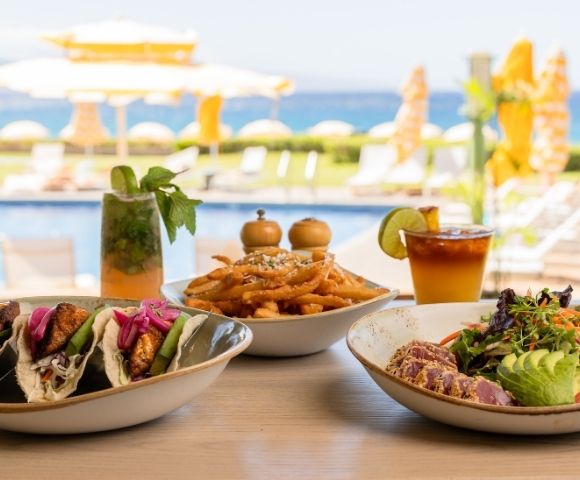 A table with fish tacos, seasoned fries, and a colorful salad. Drinks in the background; poolside setting with umbrellas and ocean view, conveying a relaxed, tropical vibe.