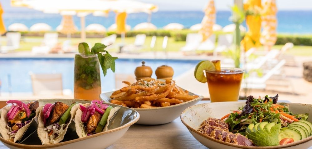 A table with fish tacos, seasoned fries, and a colorful salad. Drinks in the background; poolside setting with umbrellas and ocean view, conveying a relaxed, tropical vibe.