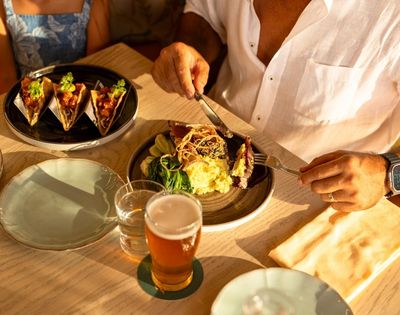 A person in a white shirt enjoys a meal at a sunlit table, featuring a gourmet dish and a glass of beer. The mood is relaxed and inviting.