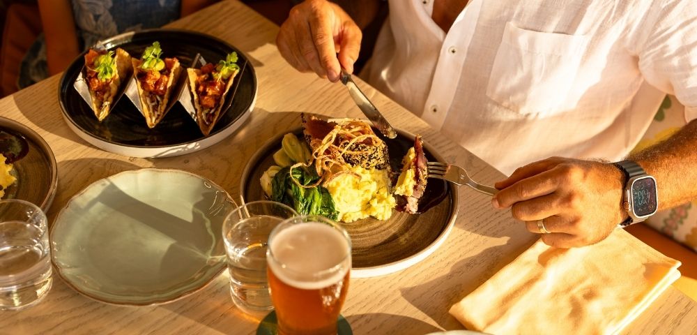 A person in a white shirt enjoys a meal at a sunlit table, featuring a gourmet dish and a glass of beer. The mood is relaxed and inviting.