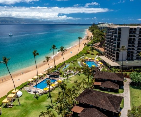 Aerial view of a tropical beach with clear blue waters and golden sand. Palm trees and resort buildings line the shore, creating a serene, vacation vibe.