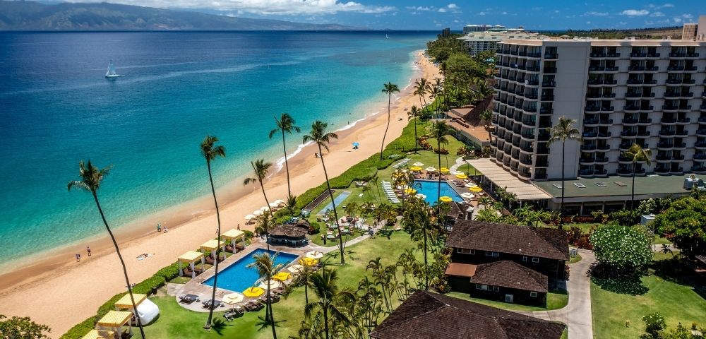 Aerial view of a tropical beach with clear blue waters and golden sand. Palm trees and resort buildings line the shore, creating a serene, vacation vibe.