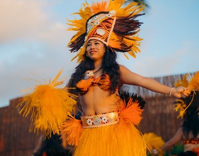 A woman performs a traditional dance in colorful attire, featuring a vibrant feathered headdress and skirt, radiating joy and energy against a blue sky.