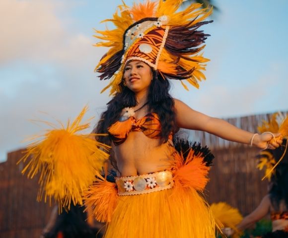 A woman performs a traditional dance in colorful attire, featuring a vibrant feathered headdress and skirt, radiating joy and energy against a blue sky.