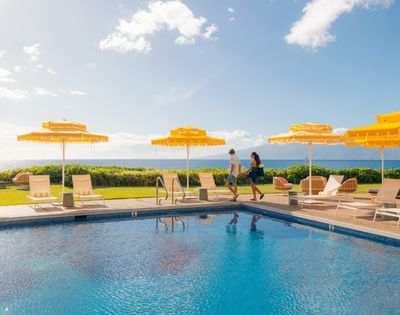 A couple walks along a sunny poolside with vibrant yellow umbrellas and lounge chairs, overlooking a lush lawn and ocean, evoking a relaxed, tropical vibe.