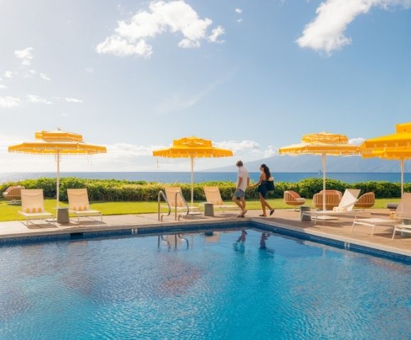 A couple walks along a sunny poolside with vibrant yellow umbrellas and lounge chairs, overlooking a lush lawn and ocean, evoking a relaxed, tropical vibe.