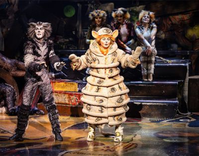 Performer in the Cats musical wearing a distinctive cream-colored costume with layered circular segments, standing center stage with arms open. Other performers in feline costumes are visible in the background near metallic stairs and props, under warm stage lighting.