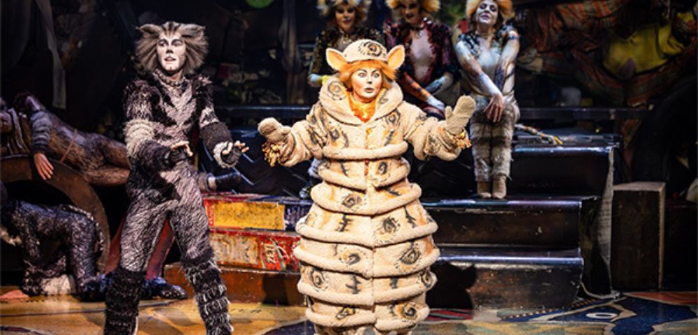 Performer in the Cats musical wearing a distinctive cream-colored costume with layered circular segments, standing center stage with arms open. Other performers in feline costumes are visible in the background near metallic stairs and props, under warm stage lighting.