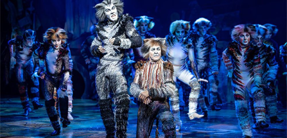 Group of performers in the Cats musical dressed in detailed cat costumes with fur textures and face paint, standing and dancing on a vibrant stage. The set includes scattered props and a backdrop with glowing blue tones, creating a mystical atmosphere.
