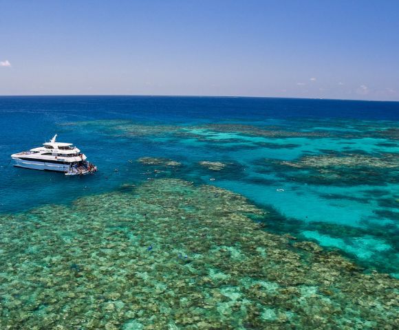 A white yacht is anchored near a vibrant coral reef in clear, turquoise ocean waters under a bright blue sky, conveying a sense of tranquility and adventure.