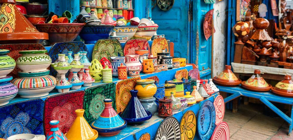 Colorful Moroccan pottery displayed in a vibrant market stall. The assortment includes tagines, bowls, and plates in bright patterns and hues, set against a blue-painted wooden backdrop with hanging decorative plates.