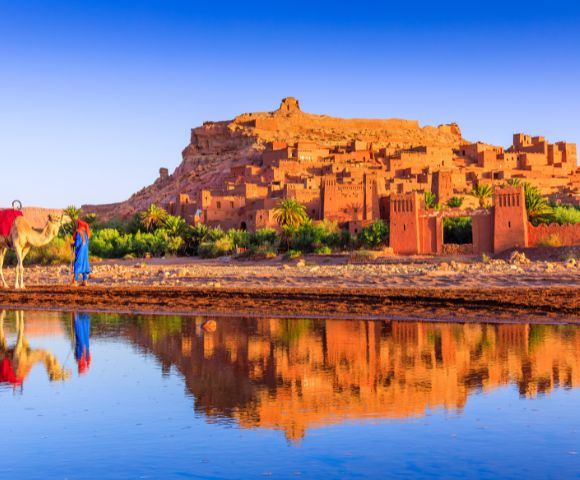 Scenic view of Ait Benhaddou, a historic fortified village in Morocco, with traditional earthen buildings glowing in warm sunlight. Two camels and their handlers walk along the riverbank, and the structures are reflected in the calm water.