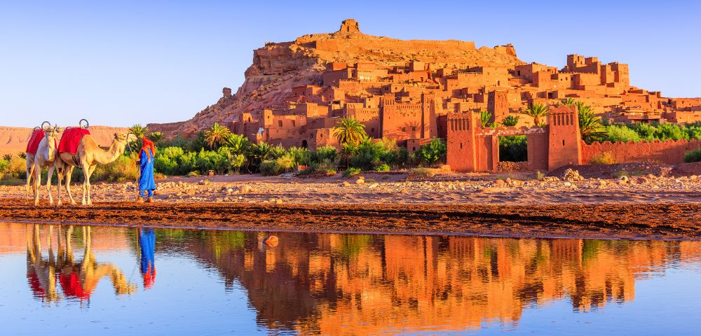Scenic view of Ait Benhaddou, a historic fortified village in Morocco, with traditional earthen buildings glowing in warm sunlight. Two camels and their handlers walk along the riverbank, and the structures are reflected in the calm water.