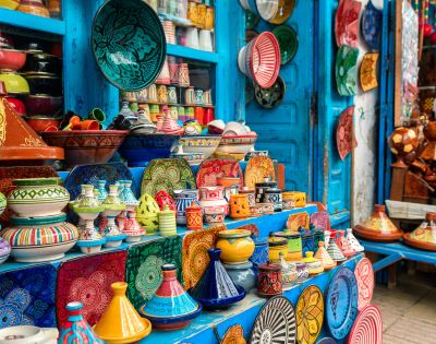 Colorful Moroccan pottery displayed in a vibrant market stall. The assortment includes tagines, bowls, and plates in bright patterns and hues, set against a blue-painted wooden backdrop with hanging decorative plates.