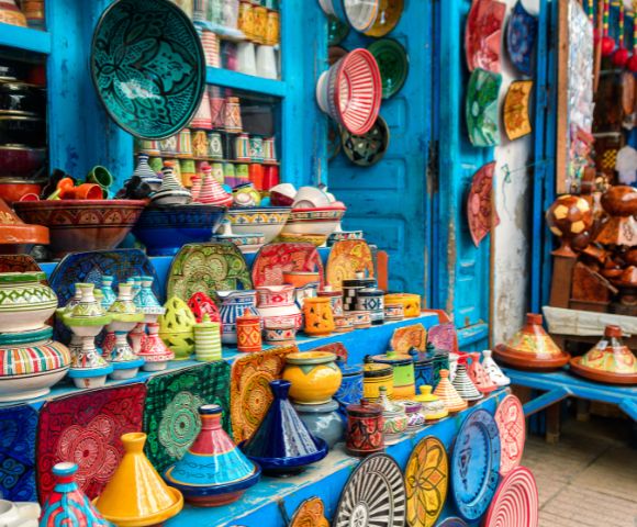 Colorful Moroccan pottery displayed in a vibrant market stall. The assortment includes tagines, bowls, and plates in bright patterns and hues, set against a blue-painted wooden backdrop with hanging decorative plates.