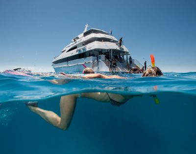 A person snorkeling in clear blue ocean water near a large white yacht. The scene is bright and sunny, evoking a sense of adventure and relaxation.