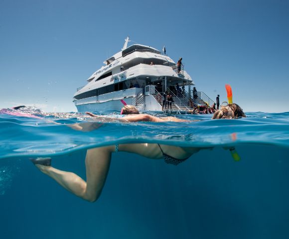 A person snorkeling in clear blue ocean water near a large white yacht. The scene is bright and sunny, evoking a sense of adventure and relaxation.
