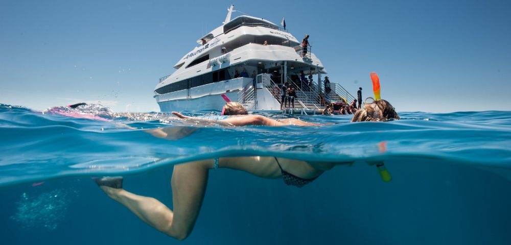 A person snorkeling in clear blue ocean water near a large white yacht. The scene is bright and sunny, evoking a sense of adventure and relaxation.