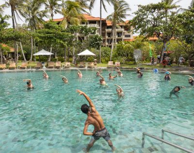 Aerial view of a resort with vibrant red-roofed buildings surrounded by lush greenery and palm trees, near a coastline with blue ocean waves.