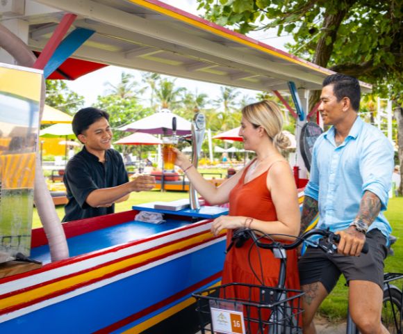 A man in a black shirt serves drinks to a smiling couple on bicycles at a colorful outdoor bar with palm trees and umbrellas in the background.