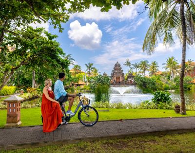 A couple rides a bicycle on a tropical path near a serene pond with a fountain, surrounded by lush greenery and palm trees under a partly cloudy sky.
