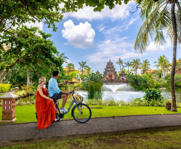 A couple rides a bicycle on a tropical path near a serene pond with a fountain, surrounded by lush greenery and palm trees under a partly cloudy sky.