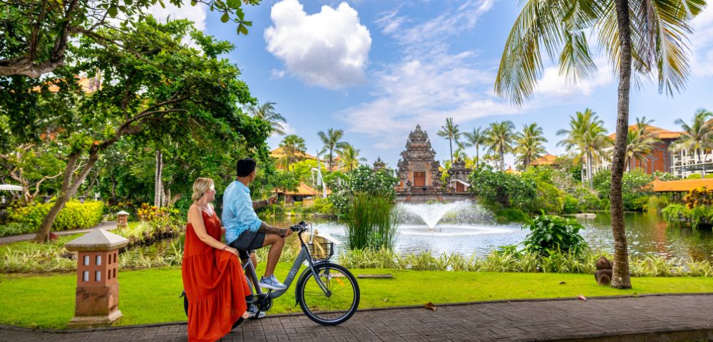 A couple rides a bicycle on a tropical path near a serene pond with a fountain, surrounded by lush greenery and palm trees under a partly cloudy sky.