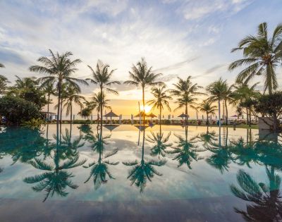 A serene sunset view with palm trees reflected in a tranquil pool, creating a peaceful and tropical ambiance. The sky is painted with soft hues of orange and blue.