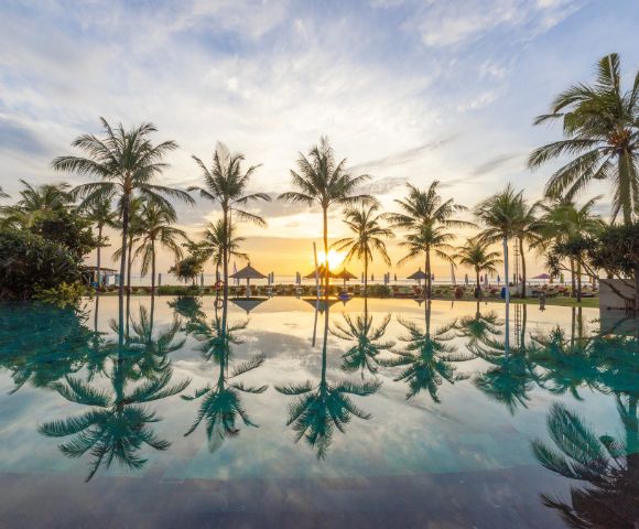 A serene sunset view with palm trees reflected in a tranquil pool, creating a peaceful and tropical ambiance. The sky is painted with soft hues of orange and blue.