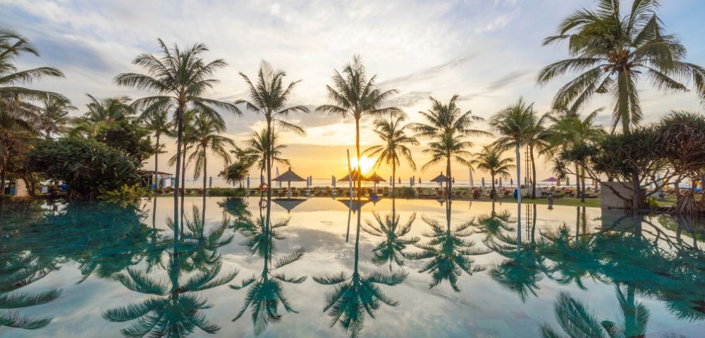 A serene sunset view with palm trees reflected in a tranquil pool, creating a peaceful and tropical ambiance. The sky is painted with soft hues of orange and blue.
