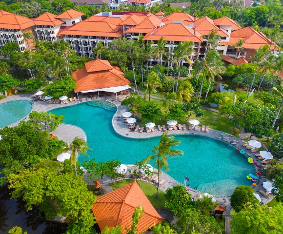 Aerial view of a luxurious resort with a large, curving turquoise pool surrounded by palm trees, deck chairs, and umbrellas. Buildings with red tile roofs are visible in the background, evoking a serene tropical atmosphere.