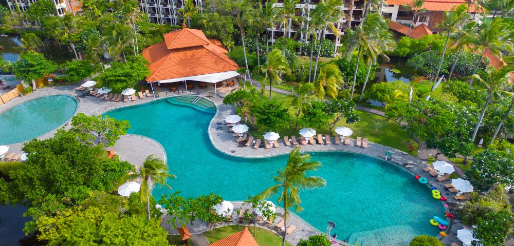 Aerial view of a luxurious resort with a large, curving turquoise pool surrounded by palm trees, deck chairs, and umbrellas. Buildings with red tile roofs are visible in the background, evoking a serene tropical atmosphere.